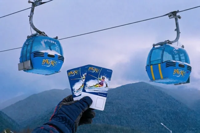 Gloved hand holding up two Bansko lift passes with blue gondola cabins overhead
