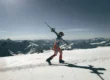 Skier walking across a snowy mountain peak holding skis over shoulders against a clear blue sky