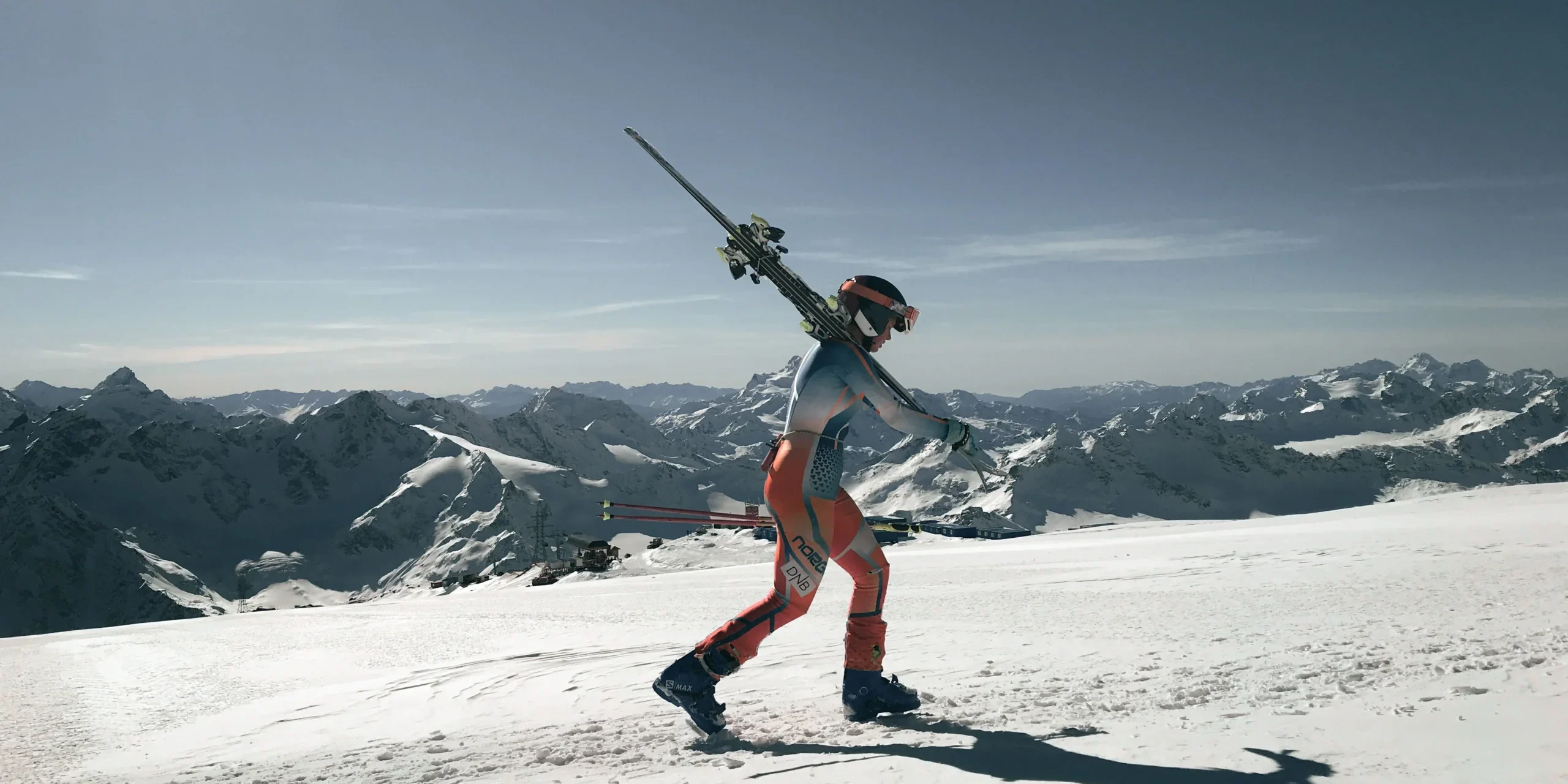 ski-fit-good-shape Skier walking across a snowy mountain peak holding skis over shoulders against a clear blue sky