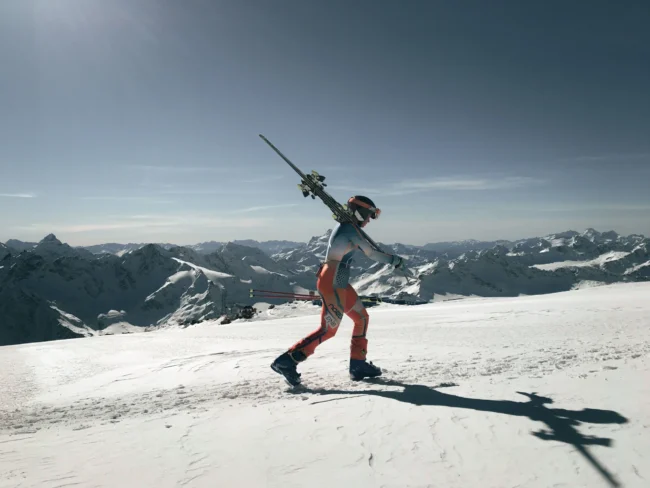 Skier walking across a snowy mountain peak holding skis over shoulders against a clear blue sky